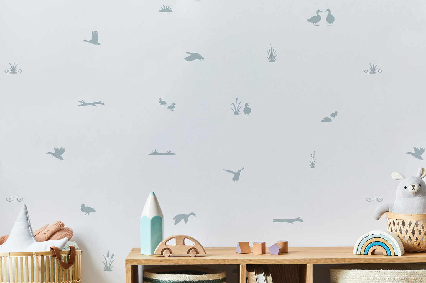 A styled nursery corner featuring light green-toned gray wall decals of ducks, ducklings, pond items and reeds on a white wall above a wooden toy shelf with toys.