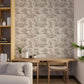 A styled interior room showing a wall covered in a beige and taupe toile-style floral wallpaper, with a wooden desk and chair, a light gray sofa, and a tall open shelving unit.