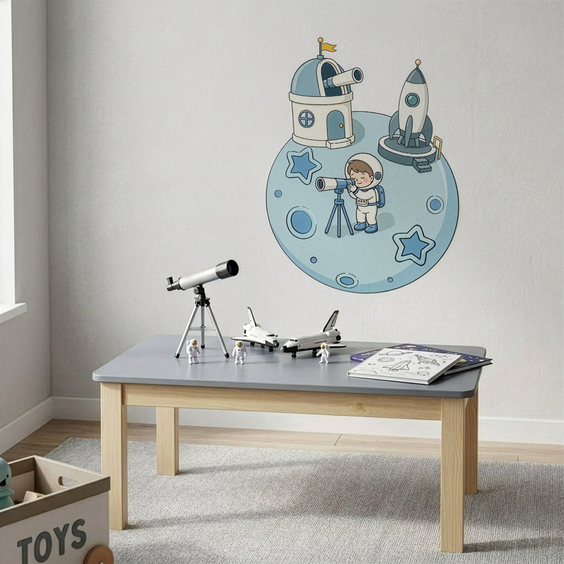 A children's room scene with a light gray wall decorated with a space-themed wall decal showing an astronaut, a moon base, and a rocket. A small gray table with wooden legs holds toy models, including a telescope, a model rocket, and an open book.