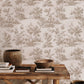 A styled interior scene showing a wall covered in beige toile wallpaper with a brown scenic landscape pattern. A rustic wooden table sits in front, styled with books, a wooden bowl, and ceramic vases.