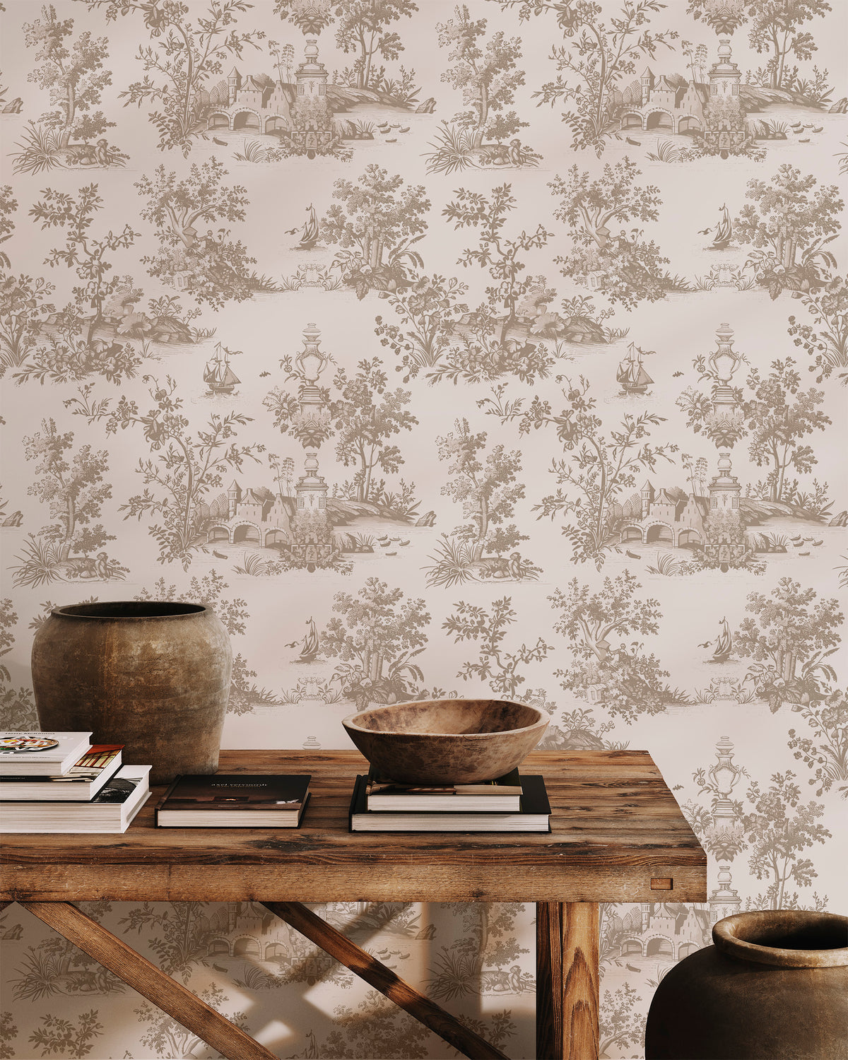 A styled interior scene showing a wall covered in beige toile wallpaper with a brown scenic landscape pattern. A rustic wooden table sits in front, styled with books, a wooden bowl, and ceramic vases.
