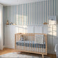 A styled nursery room featuring a light wood crib with a blue mattress and blanket, set against a white wainscoted wall with blue and white vertical stripes. A wooden dresser, white curtains, and a window are visible on the left, with warm sunlight casting shadows across the room.
