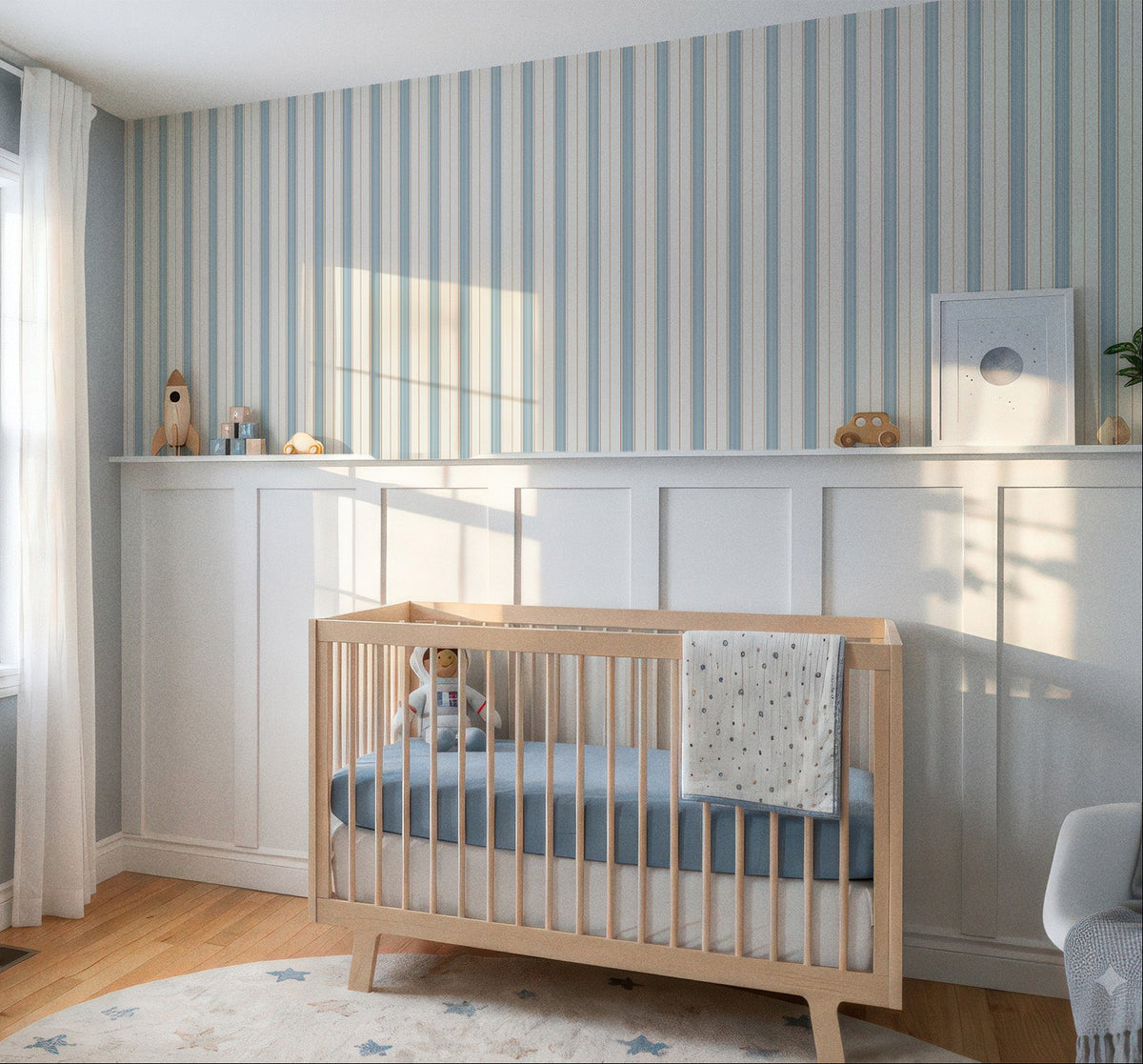 A styled nursery room featuring a light wood crib with a blue mattress and blanket, set against a white wainscoted wall with blue and white vertical stripes. A wooden dresser, white curtains, and a window are visible on the left, with warm sunlight casting shadows across the room.