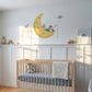 A styled nursery room featuring a light wood crib with a blue mattress, set against white wainscoting on a light gray wall. A large yellow crescent moon wall decal with a cartoon child is mounted above the crib.