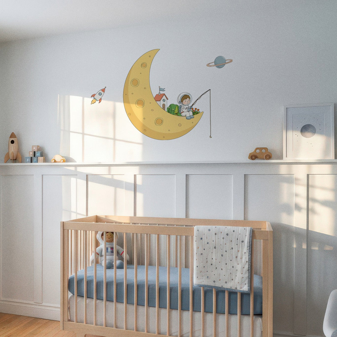 A styled nursery room featuring a light wood crib with a blue mattress, set against white wainscoting on a light gray wall. A large yellow crescent moon wall decal with a cartoon child is mounted above the crib.