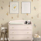 A nursery room scene featuring a pale blush pink three-drawer dresser with gold knobs, topped with a changing pad and small accessories. The dresser is set against a warm beige wall decorated with whimsical goose decals and two framed prints, with a small stool on the left and a plush toy basket on the right.