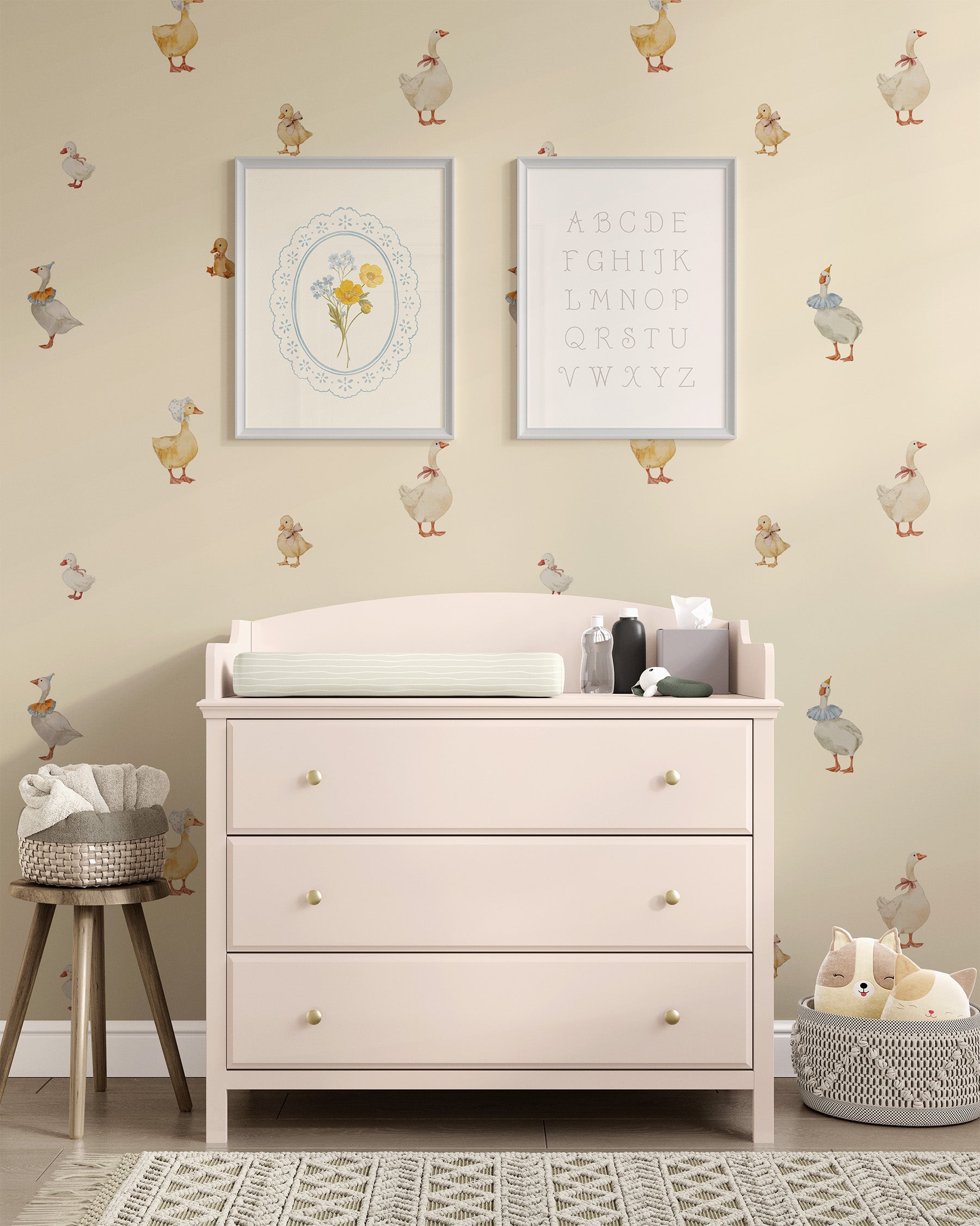A nursery room scene featuring a pale blush pink three-drawer dresser with gold knobs, topped with a changing pad and small accessories. The dresser is set against a warm beige wall decorated with whimsical goose decals and two framed prints, with a small stool on the left and a plush toy basket on the right.
