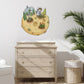 A whimsical wall decal of an astronaut gardening on a moon is displayed on a white wall above a light wood dresser in a styled nursery room setting.