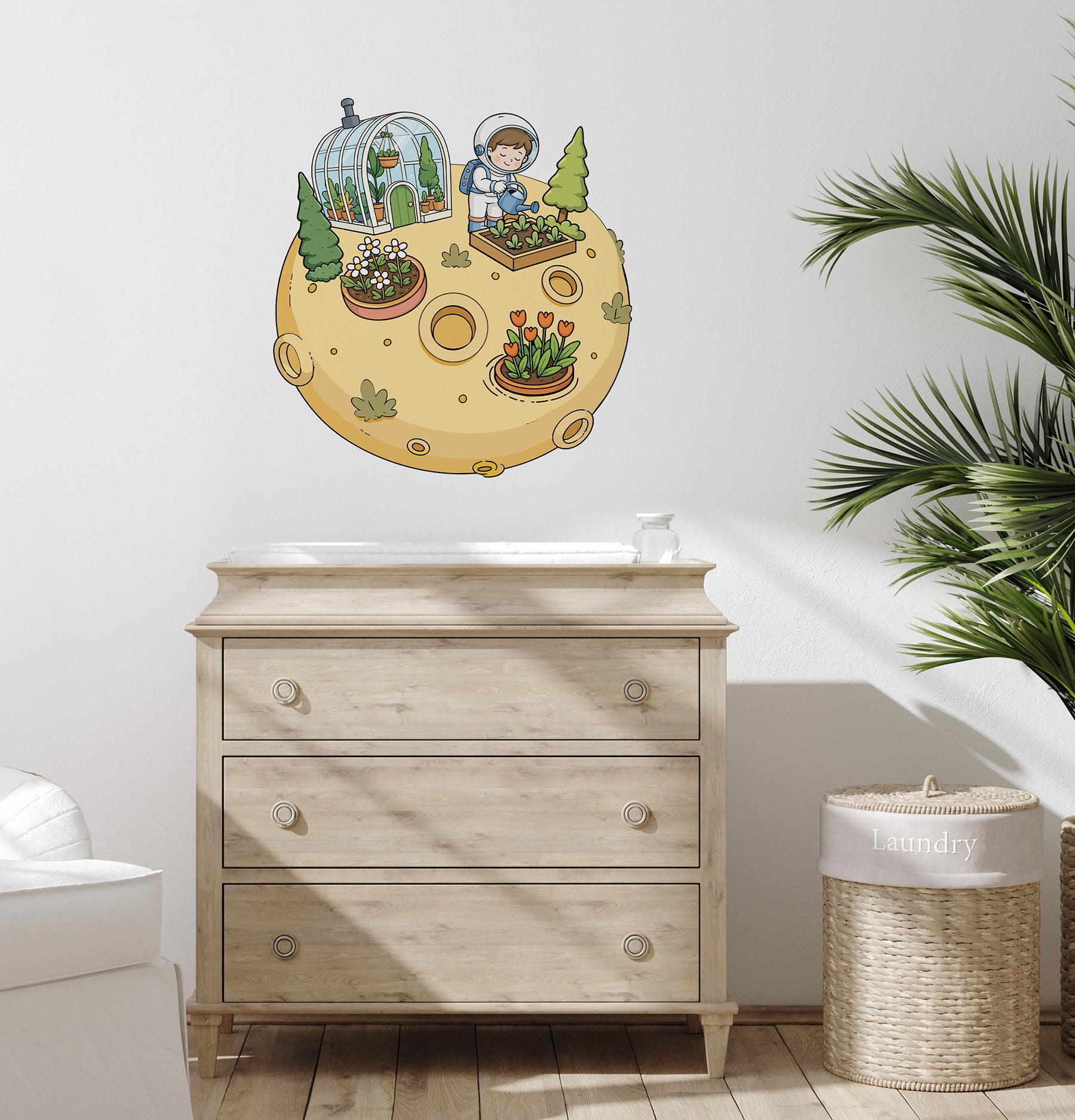 A whimsical wall decal of an astronaut gardening on a moon is displayed on a white wall above a light wood dresser in a styled nursery room setting.
