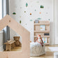 A styled children's room corner with a large house-shaped wooden play structure in the foreground, a woven pouf with a plush toy, a small wooden shelf, and fishing and camping themed wall decals on a white wall.