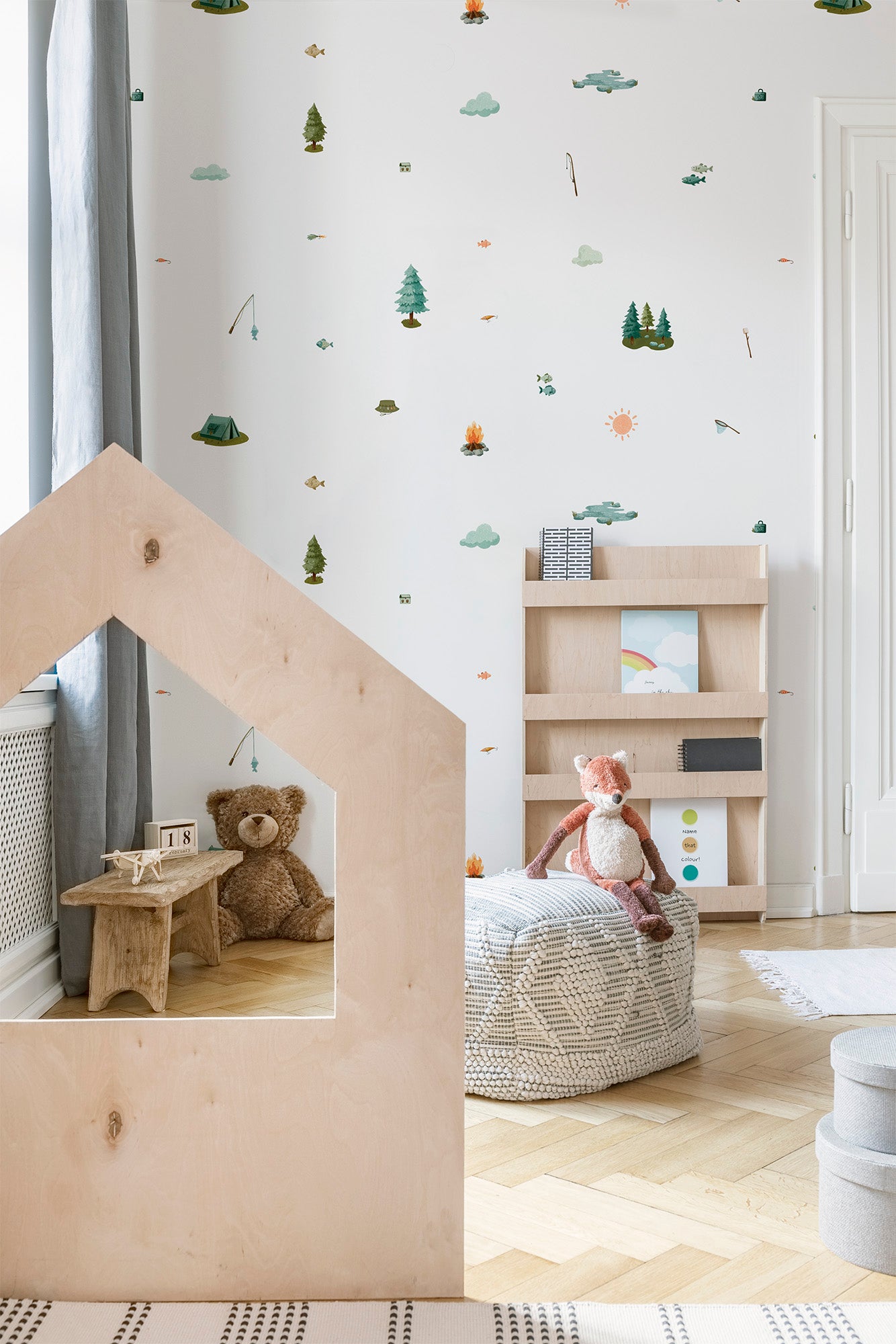 A styled children's room corner with a large house-shaped wooden play structure in the foreground, a woven pouf with a plush toy, a small wooden shelf, and fishing and camping themed wall decals on a white wall.