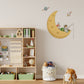 A children's room scene with a large wall decal featuring a yellow crescent moon, a child astronaut fishing, a small rocket and planet. The room includes a wooden toy shelf with books and toys, a white storage bin, and a small stool with a plush toy.