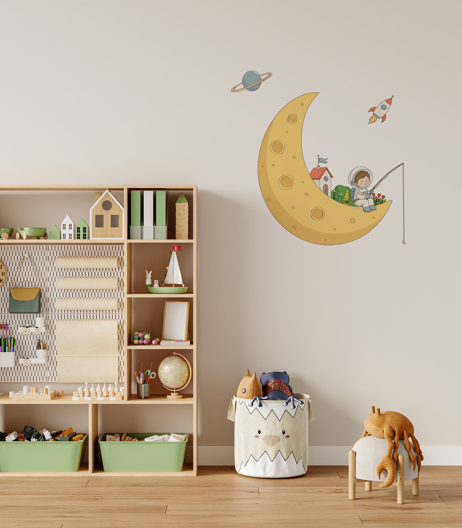 A children's room scene with a large wall decal featuring a yellow crescent moon, a child astronaut fishing, a small rocket and planet. The room includes a wooden toy shelf with books and toys, a white storage bin, and a small stool with a plush toy.