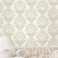 Vintage style damask wallpaper featuring ribbons and florals in a light sage green and cream pattern, shown on a wall above a white crib with a soft white blanket and a white bedside table with a lamp.