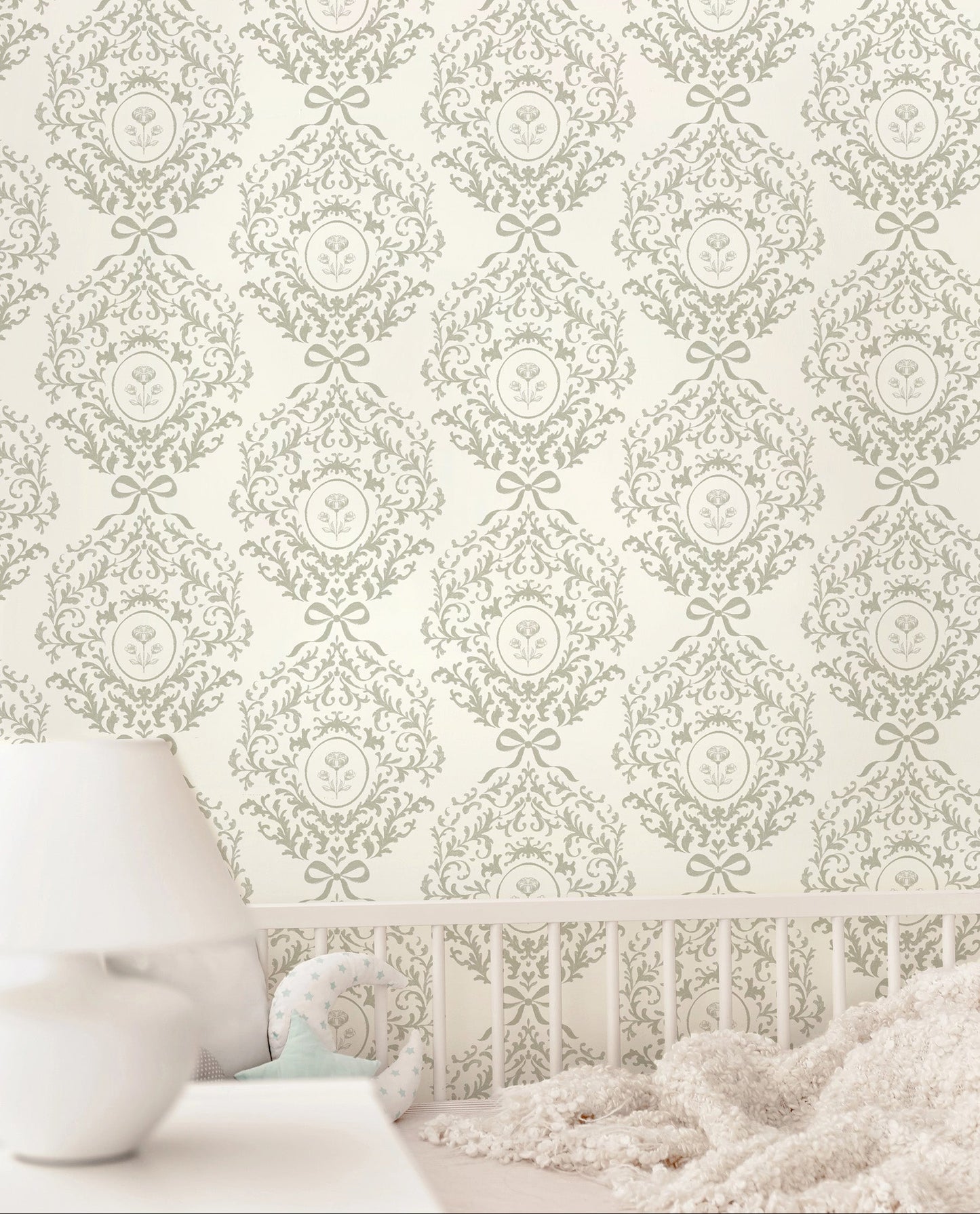 Vintage style damask wallpaper featuring ribbons and florals in a light sage green and cream pattern, shown on a wall above a white crib with a soft white blanket and a white bedside table with a lamp.
