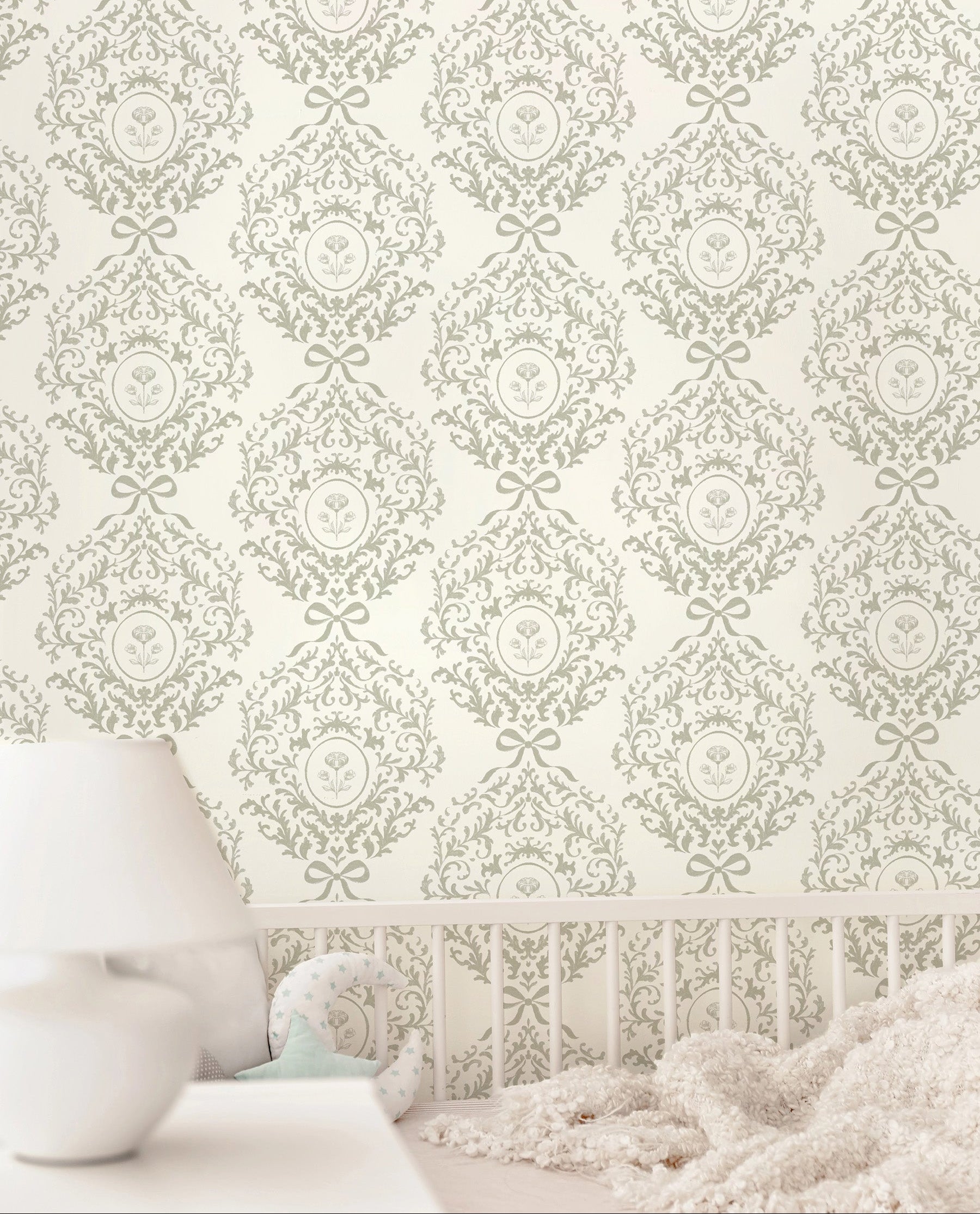 Vintage style damask wallpaper featuring ribbons and florals in a light sage green and cream pattern, shown on a wall above a white crib with a soft white blanket and a white bedside table with a lamp.