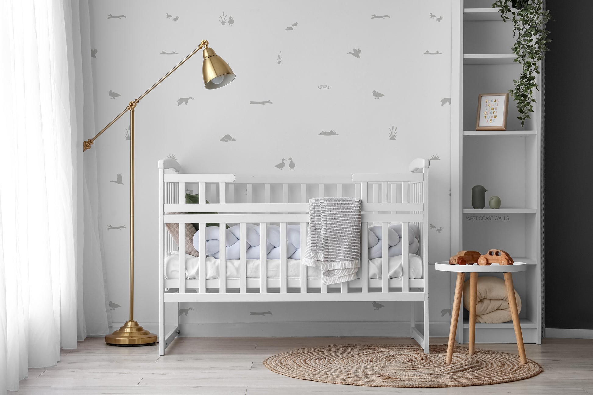 A styled nursery interior featuring subtle grey duck pond themed wall decals behind a white wooden crib with white bedding. 