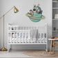 A wall decal of a cartoon astronaut sitting on a teal-blue music themed planet while playing guitar. It is displayed on a white wall above a white wooden baby crib in a styled nursery room.
