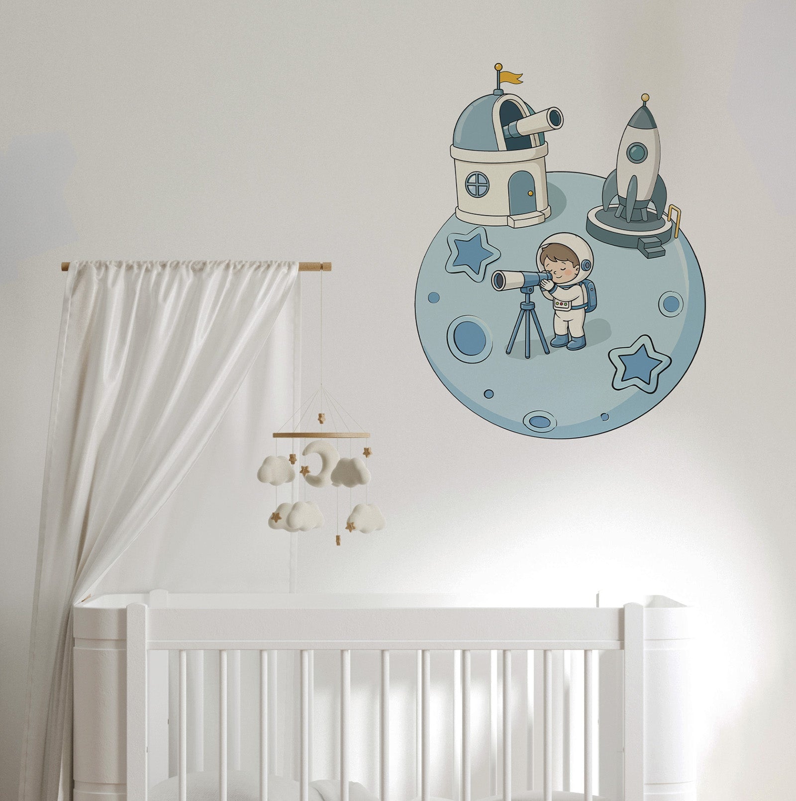 A nursery wall decal featuring a cartoon astronaut, space station, and rocket on a blue moon, shown above a white crib with a white canopy and cloud mobile.
