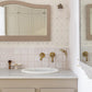 A styled bathroom vanity featuring a cream-colored countertop, a white sink basin, brass wall-mounted faucets, and a beige cabinet with brass knobs. The background showcases a light pink tiled backsplash, a soft blue diamond patterned wallpaper, and a matching arched mirror with a beige frame.
