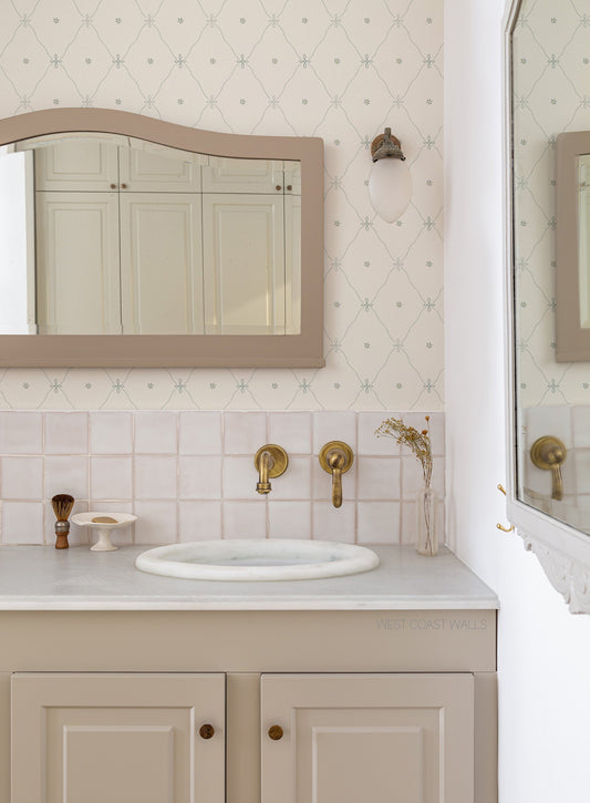 A styled bathroom vanity featuring a cream-colored countertop, a white sink basin, brass wall-mounted faucets, and a beige cabinet with brass knobs. The background showcases a light pink tiled backsplash, a soft blue diamond patterned wallpaper, and a matching arched mirror with a beige frame.