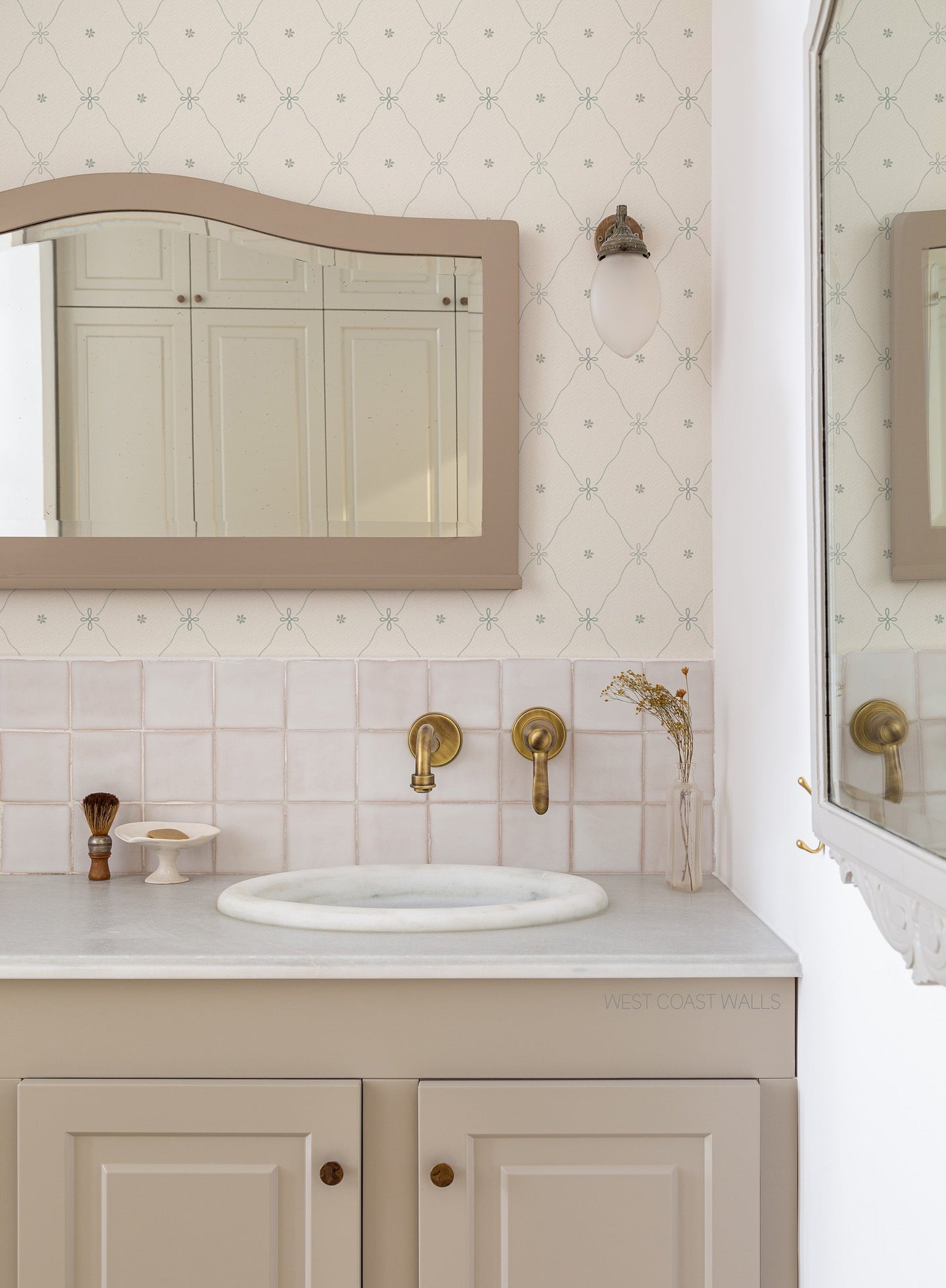 A styled bathroom vanity featuring a cream-colored countertop, a white sink basin, brass wall-mounted faucets, and a beige cabinet with brass knobs. The background showcases a light pink tiled backsplash, a soft blue diamond patterned wallpaper, and a matching arched mirror with a beige frame.