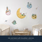 A set of whimsical space-themed wall decals displayed on a light gray wall above a wooden table and two cribs. The decals include a large yellow crescent moon with an astronaut fishing, several small planets with houses, a rocket ship, and a satellite, with text at the bottom reading 'Mix and Match with Moonfisher collection'. Additional planet decals are not included in the moonfisher set