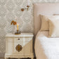A styled bedroom corner featuring a vintage-style white nightstand with ornate brass hardware, a lit candle, and a beige upholstered bed with cream bedding, set against a green and white damask wallpaper wall.