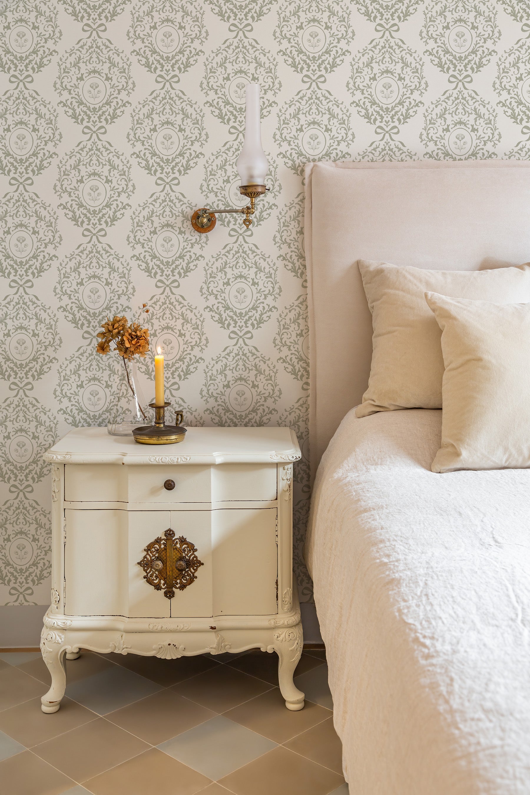 A styled bedroom corner featuring a vintage-style white nightstand with ornate brass hardware, a lit candle, and a beige upholstered bed with cream bedding, set against a green and white damask wallpaper wall.